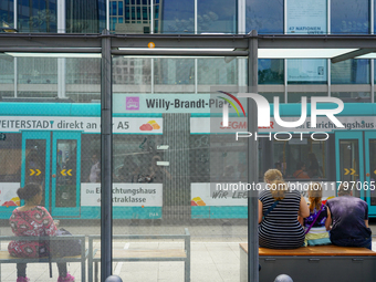 Passengers sit on benches at a tram stop near Willy-Brandt-Platz in a modern city setting on August 15, 2023. Through the glass panels, a tu...