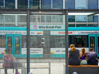 Passengers sit on benches at a tram stop near Willy-Brandt-Platz in a modern city setting on August 15, 2023. Through the glass panels, a tu...