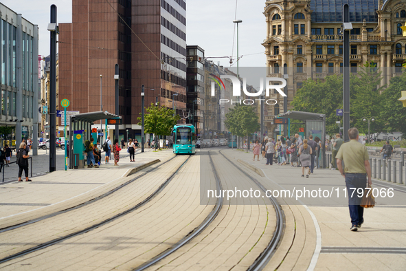 A turquoise tram approaches the bustling tram stop in the city on August 15, 2023, where passengers gather to board. The scene captures the...