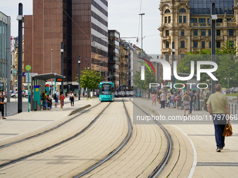 A turquoise tram approaches the bustling tram stop in the city on August 15, 2023, where passengers gather to board. The scene captures the...