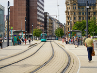 A turquoise tram approaches the bustling tram stop in the city on August 15, 2023, where passengers gather to board. The scene captures the...