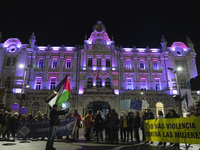 Demonstrators carry signs saying no more violence against women and a Palestinian flag as they walk past the town hall, which is lit up in p...