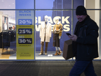 A man carries a box and a smartphone as he walks past a shop window with Black Week sales displayed ahead of Black Friday in Warsaw, Poland,...