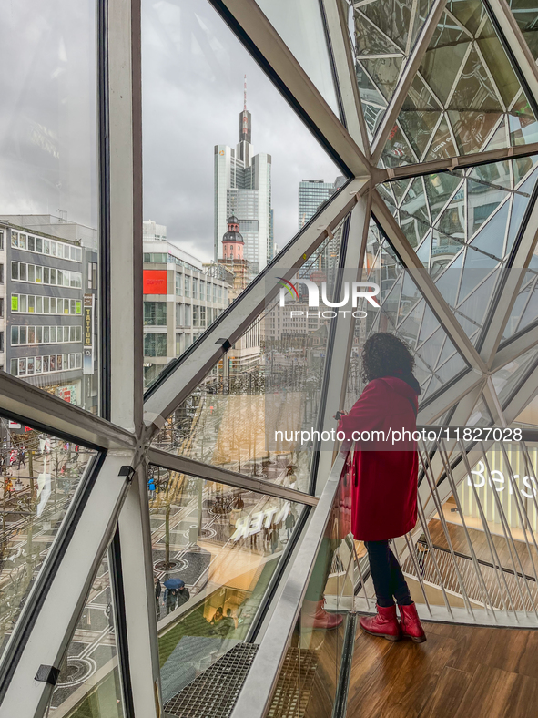 A woman in a red coat stands inside a shopping mall with a geometric glass window overlooking the Frankfurt am Main skyline, dominated by th...