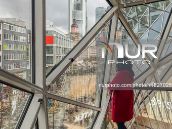 A woman in a red coat stands inside a shopping mall with a geometric glass window overlooking the Frankfurt am Main skyline, dominated by th...