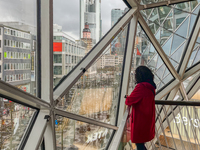 A woman in a red coat stands inside a shopping mall with a geometric glass window overlooking the Frankfurt am Main skyline, dominated by th...