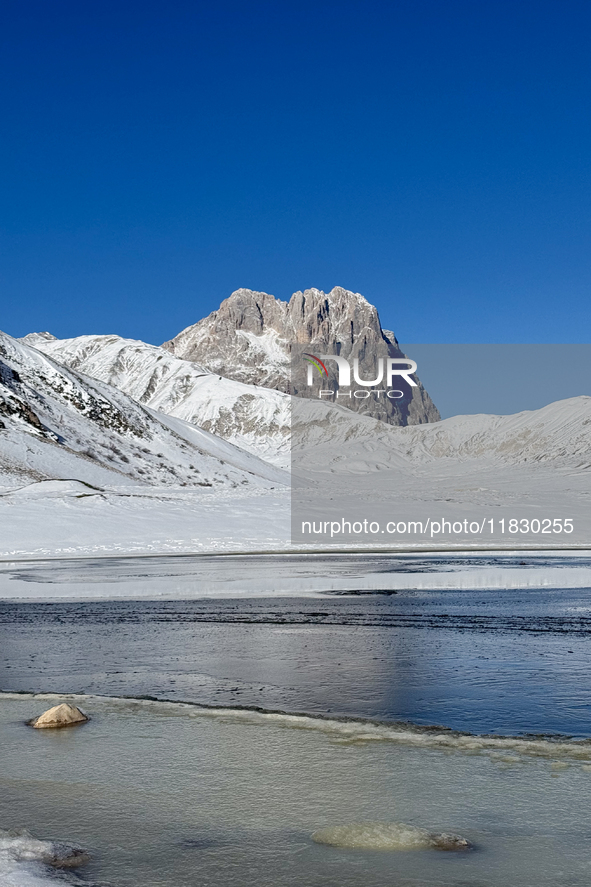 The frozen surface of a mountain lake reflects the Corno Grande under a blue sky in Campo Imperatore, Gran Sasso d'Italia, Italy, on Novembe...