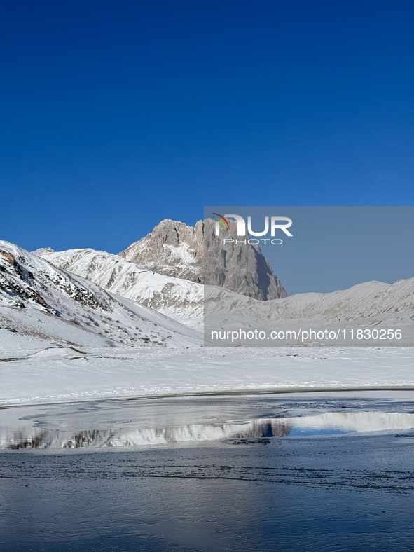 The frozen surface of a mountain lake reflects the Corno Grande under a blue sky in Campo Imperatore, Gran Sasso d'Italia, Italy, on Novembe...