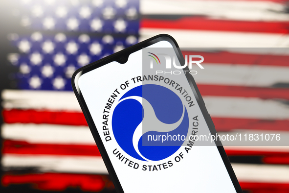 The United States Department of Transportation (USDOT or DOT) logo appears on the screen of a smartphone, and in the background, the flag of...