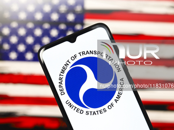 The United States Department of Transportation (USDOT or DOT) logo appears on the screen of a smartphone, and in the background, the flag of...