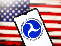 The United States Department of Transportation (USDOT or DOT) logo appears on the screen of a smartphone, and in the background, the flag of...