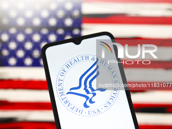 The United States Department of Health and Human Services (HHS) logo appears on the screen of a smartphone, and in the background, the flag...