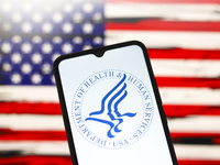 The United States Department of Health and Human Services (HHS) logo appears on the screen of a smartphone, and in the background, the flag...