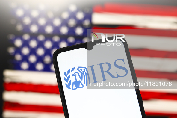 The Internal Revenue Service (IRS) logo appears on the screen of a smartphone, and in the background, the flag of the United States is on th...