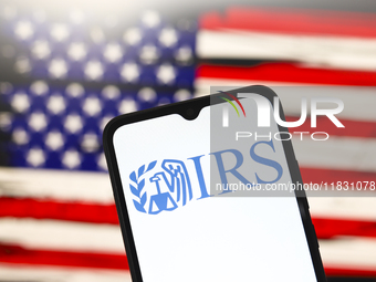 The Internal Revenue Service (IRS) logo appears on the screen of a smartphone, and in the background, the flag of the United States is on th...