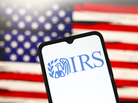 The Internal Revenue Service (IRS) logo appears on the screen of a smartphone, and in the background, the flag of the United States is on th...