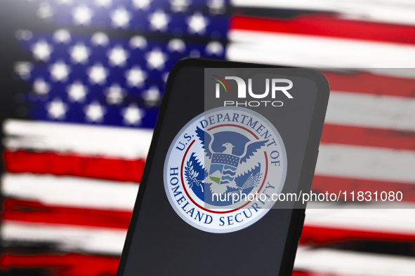 The United States Department of Homeland Security (DHS) logo appears on the screen of a smartphone, and in the background, the flag of the U...