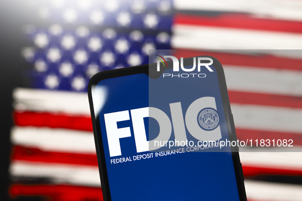 The United States Federal Deposit Insurance Corporation (FDIC) logo appears on the screen of a smartphone, and in the background, the flag o...