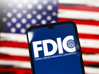 The United States Federal Deposit Insurance Corporation (FDIC) logo appears on the screen of a smartphone, and in the background, the flag o...