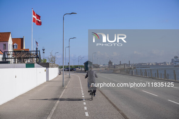 A woman cycles on a bicycle along a street running along the port quay with a Danish flag on her right hand in Nordby, Fano Island, Denmark,...