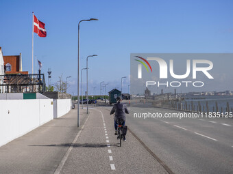 A woman cycles on a bicycle along a street running along the port quay with a Danish flag on her right hand in Nordby, Fano Island, Denmark,...