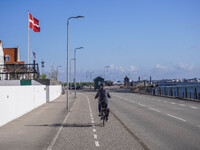 A woman cycles on a bicycle along a street running along the port quay with a Danish flag on her right hand in Nordby, Fano Island, Denmark,...