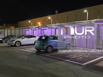 This nighttime image shows an electric vehicle charging station in an urban setting. The charging points are equipped with various models of...