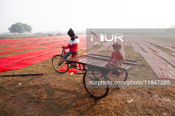 A young child sits on a rickshaw van while red textile strips dry in the sun, spread across an open field near Narsingdi, Dhaka, Bangladesh,...