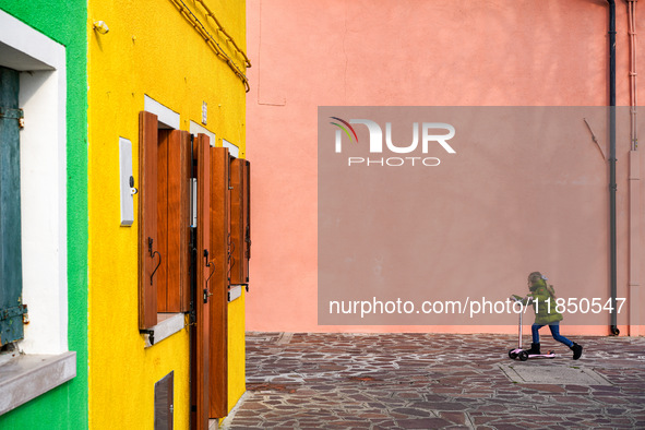 In Burano, Italy, on November 13, 2021, a child in a green jacket and jeans rides a pink scooter past vivid yellow and pink walls with woode...