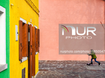 In Burano, Italy, on November 13, 2021, a child in a green jacket and jeans rides a pink scooter past vivid yellow and pink walls with woode...