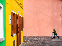 In Burano, Italy, on November 13, 2021, a child in a green jacket and jeans rides a pink scooter past vivid yellow and pink walls with woode...