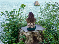 A woman sits alone on a tree stump surrounded by lush green plants, gazing out over the calm waters of the lake at Starnberger See in Starnb...