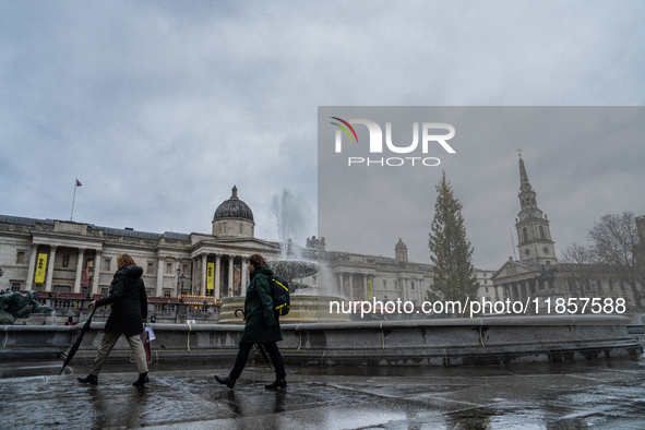 People walk past the fountains at Trafalgar Square under cloudy skies during Storm Darragh on December 8, 2024, in London, United Kingdom. L...