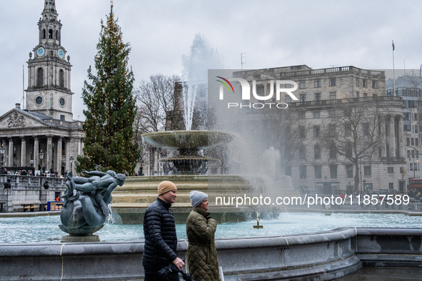 People walk past the fountains at Trafalgar Square under cloudy skies during Storm Darragh on December 8, 2024, in London, United Kingdom. L...