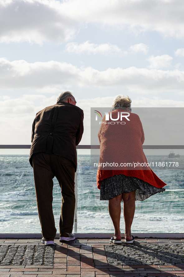 An elderly senior couple stands by a railing on a promenade in windy weather and looks out to sea in Westerland, Sylt, Germany. 