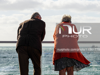 An elderly senior couple stands by a railing on a promenade in windy weather and looks out to sea in Westerland, Sylt, Germany. (