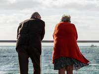 An elderly senior couple stands by a railing on a promenade in windy weather and looks out to sea in Westerland, Sylt, Germany. (