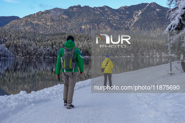 Two hikers, one wearing a green jacket and a backpack, the other in a bright yellow coat, walk along the snow-covered trail beside Lake Eibs...