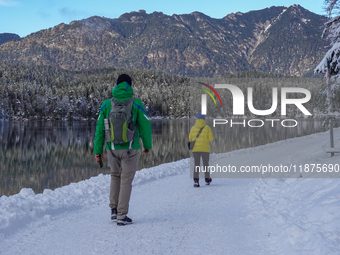 Two hikers, one wearing a green jacket and a backpack, the other in a bright yellow coat, walk along the snow-covered trail beside Lake Eibs...