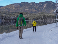 Two hikers, one wearing a green jacket and a backpack, the other in a bright yellow coat, walk along the snow-covered trail beside Lake Eibs...