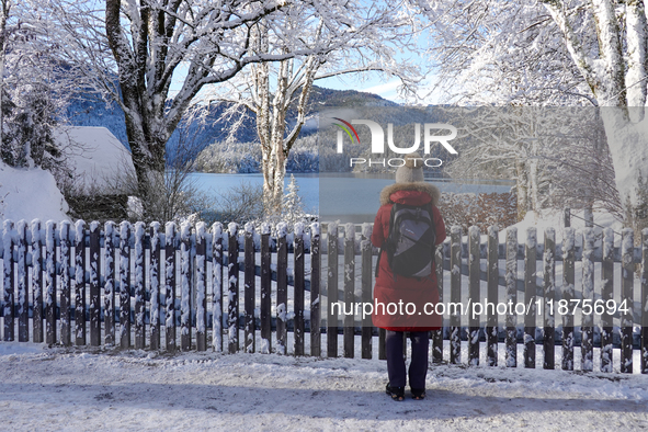 A hiker stands quietly near a wooden fence covered in fresh snow, gazing out over the calm and reflective Lake Eibsee in Grainau, Bavaria, G...