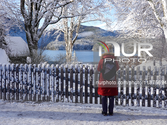 A hiker stands quietly near a wooden fence covered in fresh snow, gazing out over the calm and reflective Lake Eibsee in Grainau, Bavaria, G...