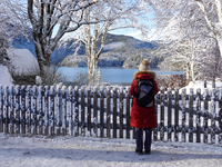 A hiker stands quietly near a wooden fence covered in fresh snow, gazing out over the calm and reflective Lake Eibsee in Grainau, Bavaria, G...