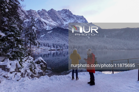 Two visitors stand on the snowy banks of Lake Eibsee in Grainau, Bavaria, Germany, surrounded by a winter landscape. One person, wearing a b...