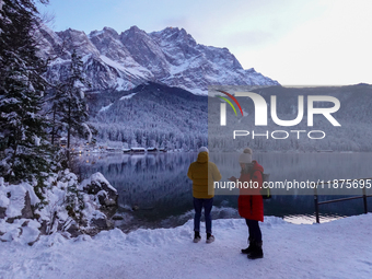 Two visitors stand on the snowy banks of Lake Eibsee in Grainau, Bavaria, Germany, surrounded by a winter landscape. One person, wearing a b...