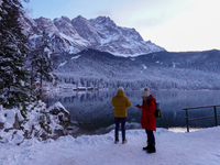 Two visitors stand on the snowy banks of Lake Eibsee in Grainau, Bavaria, Germany, surrounded by a winter landscape. One person, wearing a b...