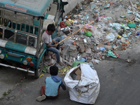 Two young child laborers collect plastic bottles and other reusable items from a roadside garbage disposal area while a worker and two schoo...