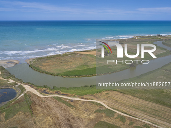 An aerial view shows the Ofanto River meeting the Adriatic Sea, highlighting the contrast between the green riverbanks and the blue sea in P...