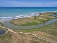 An aerial view shows the Ofanto River meeting the Adriatic Sea, highlighting the contrast between the green riverbanks and the blue sea in P...