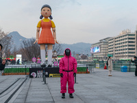 A person dressed as a ''Squid Game'' game guard stands while people take photos in front of Young-hee, a robotic doll from Netflix's ''Squid...